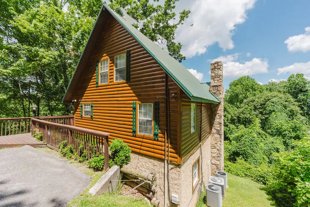 An exterior view of the log cabin, featuring a welcoming deck area, green roof, and stone chimney. The inviting outdoor space hints at possibilities for group gatherings and relaxation, while the cabin's structure promises comfort within a cozy mountain retreat.