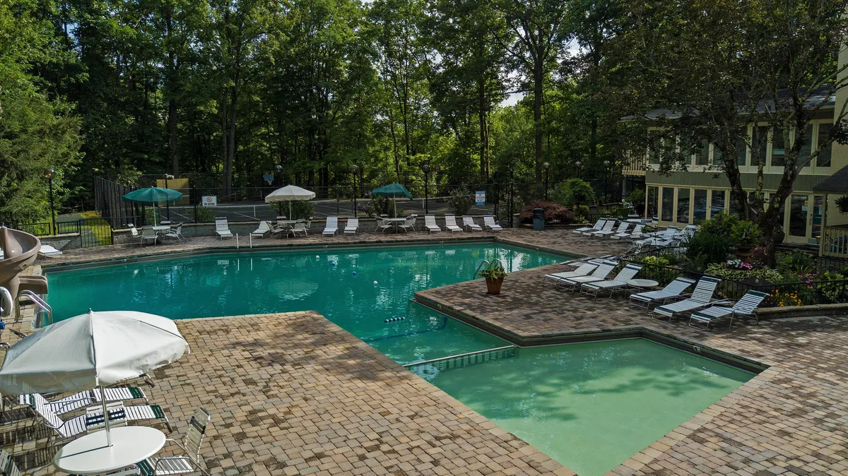 This image features a sparkling swimming pool surrounded by lounge chairs and umbrellas, with trees and a patio area providing a relaxing and inviting outdoor setting. The scene conveys an atmosphere of fun, family-friendly recreation, and relaxation in the heart of nature.