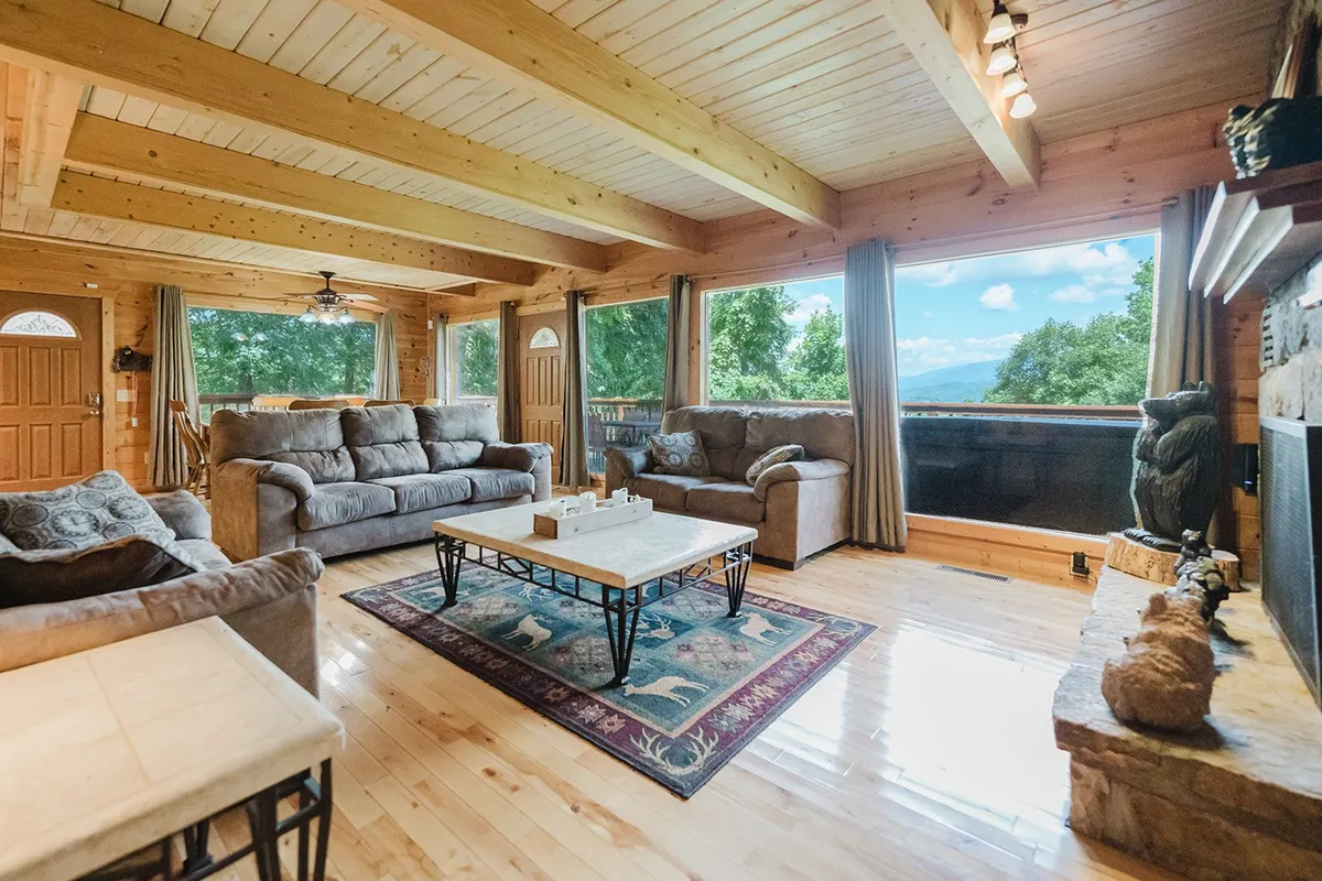 A warm, spacious living area featuring comfortable sofas arranged around a fireplace. The room is accented with wooden beams and ceiling, a central coffee table, and expansive windows that reveal scenic outdoor views. Sunlight pours in, creating an inviting atmosphere ideal for gathering and relaxing.