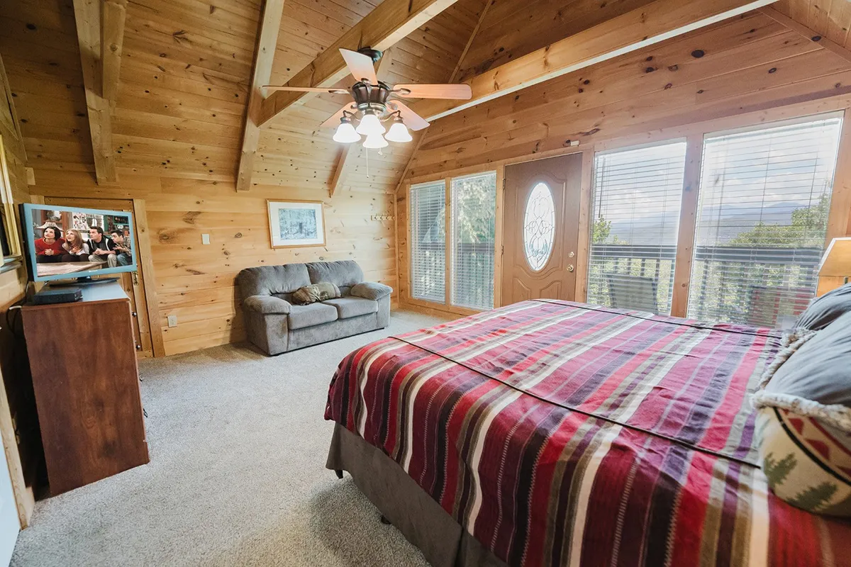 A comfortable bedroom featuring a king-sized bed with striped bedding, a sofa for lounging, large windows that invite natural light, and a TV for entertainment, creating a welcoming space for relaxation and privacy.