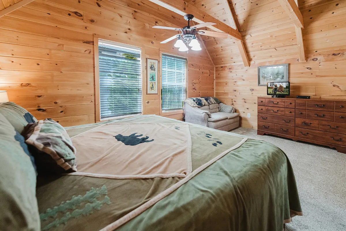 A tranquil king bedroom filled with morning sunlight, featuring a king-sized bed with a patterned blanket, a wooden dresser, a comfortable armchair by large windows with blinds, and cozy, welcoming decor.