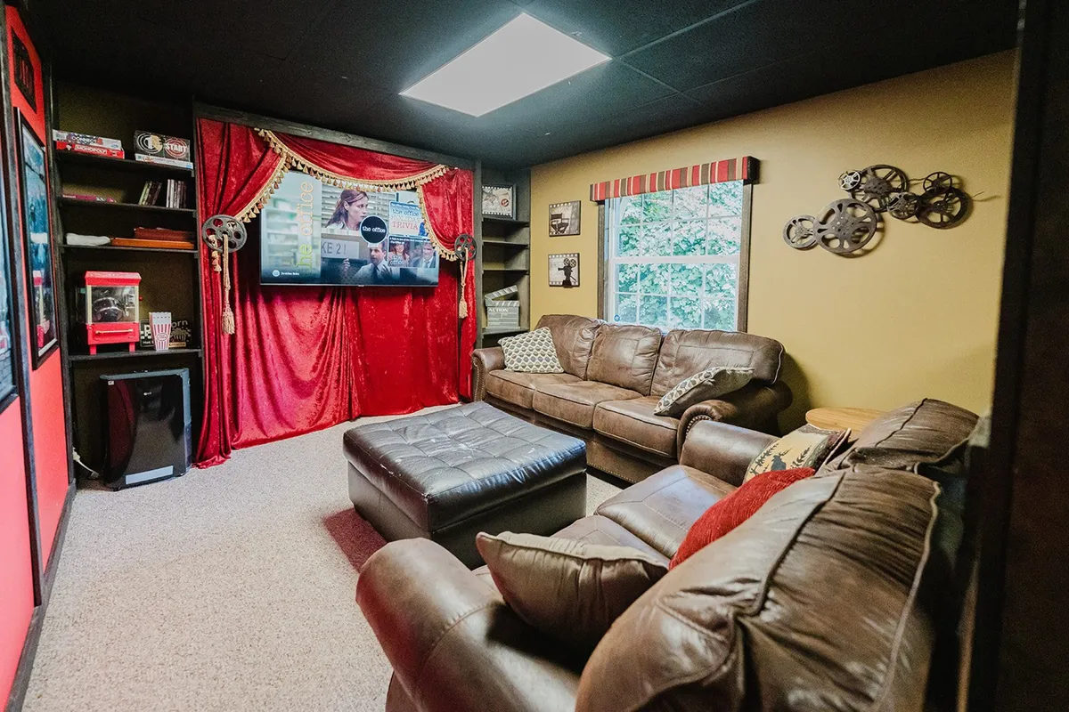 A cozy theatre lounge setup with a large TV framed by rich red curtains, comfortable leather sofas, decorative wall art, and bookshelves with memorabilia. The space is tailored for movie nights, themed watch parties, and group relaxation.