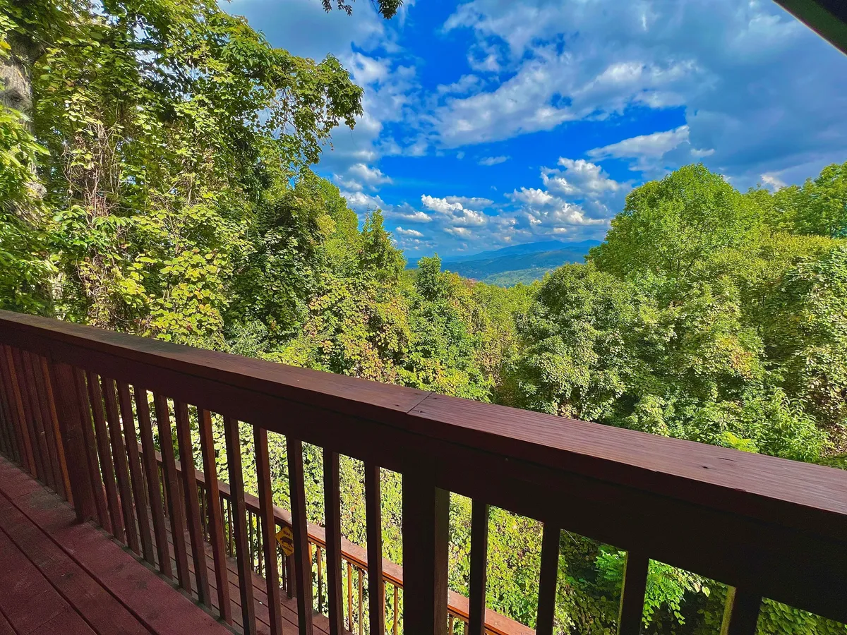 A scenic view from a wooden deck features a sturdy railing, lush greenery in the foreground, and rolling, distant mountains beneath a blue sky. The deck appears inviting for morning coffee, birdwatching, or simply soaking in the tranquil Smokies landscape.