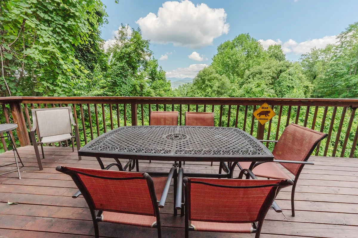 An exterior dining table set on a deck is surrounded by rich greenery, with distant mountains and a dramatic, cloudy blue sky in the background. The setup suggests opportunities for al fresco dining, morning coffee, or evening relaxation amid the natural setting.