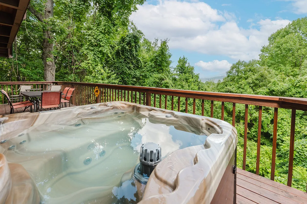 This image features a bubbling hot tub set on a spacious wooden deck, surrounded by lush greenery and offering glimpses of distant mountain views. The inviting setup suggests relaxation and social gathering, with the deck seamlessly blending into the natural forest setting.