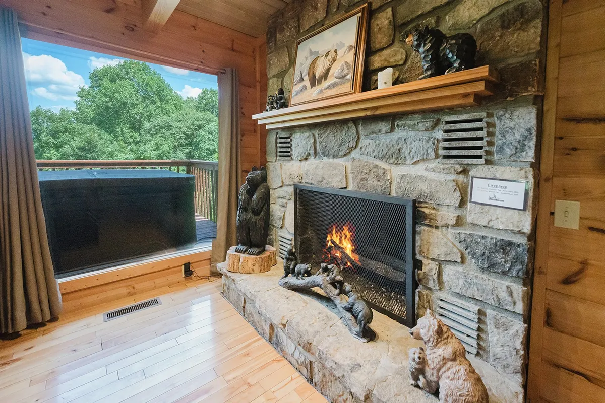 A cozy seating area centered around a striking stone fireplace with a wooden mantel, framed by a large window offering scenic views. Decorative animal sculptures accent the room, and natural light bathes the inviting space, blending interior comfort with the surrounding landscape.