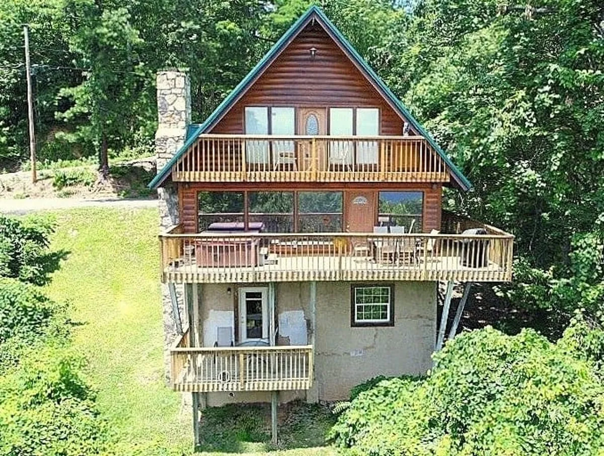 A cozy wooden balcony with large windows and a stone chimney, set amidst a forested backdrop. The balcony suggests opportunities for outdoor dining, stargazing, or simply soaking in the serenity of the Smoky Mountains while staying close to the comforts of the cabin.