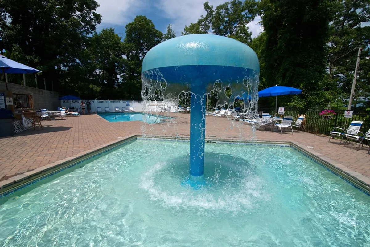 A lively resort area with a water fountain, swimming pool, lounge chairs, and umbrellas—inviting for group swimming, sunbathing, and playful afternoons. The scene conveys opportunities for communal fun and relaxation, enhancing the resort experience for all ages.