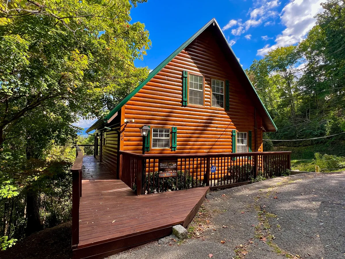 A charming log cabin exterior featuring green shutters and a wide wooden deck, surrounded by trees. The inviting entrance showcases the promise of adventure and comfort, seamlessly connecting indoor amenities with outdoor exploration.